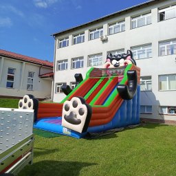 AMW Zjeżdżalnie Dmuchane - Kolorowa, dmuchana zjeżdżalnia z motywem kreskówkowym na trawie przed budynkiem. Widoczny fragment przyczepy transportowej.