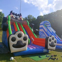 AMW Zjeżdżalnie Dmuchane - Kolorowy dmuchany plac zabaw z motywem zwierzęcym, dzieci w trakcie zabawy, buty pozostawione obok, w tle zieleń drzew i błękitne niebo. Słoneczny dzień.