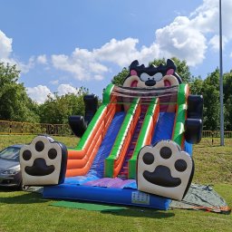 AMW Zjeżdżalnie Dmuchane - Kolorowa dmuchana zjeżdżalnia z motywem kreskówkowym na trawie, widok z dołu, błękitne niebo z chmurami w tle, częściowo widoczny samochód po lewej stronie.