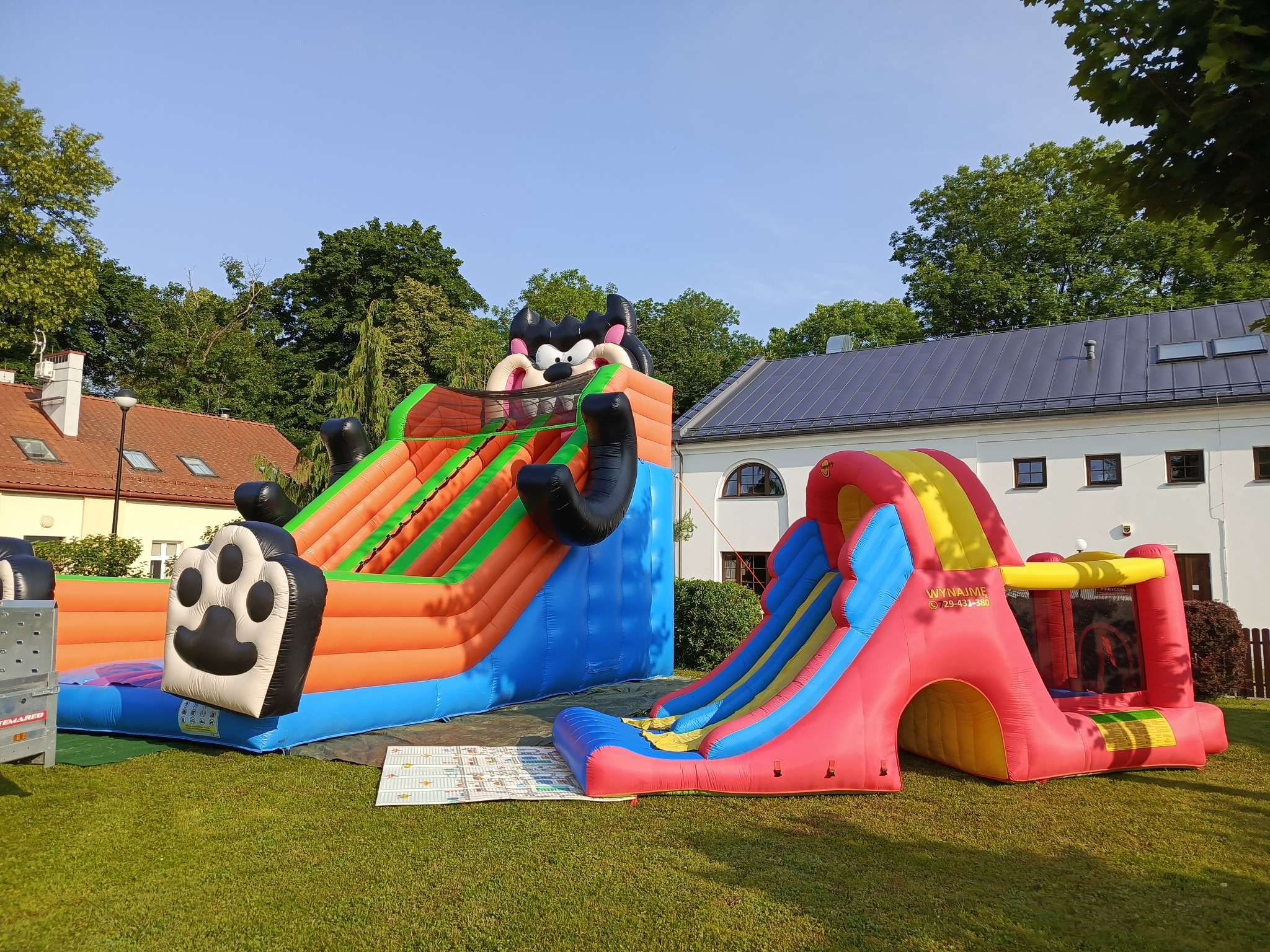 Dwa kolorowe dmuchańce na trawie: jeden z motywem kreskówkowym, drugi zjeżdżalnia z tunelem. W tle budynek z szarą dachówką i drzewa.