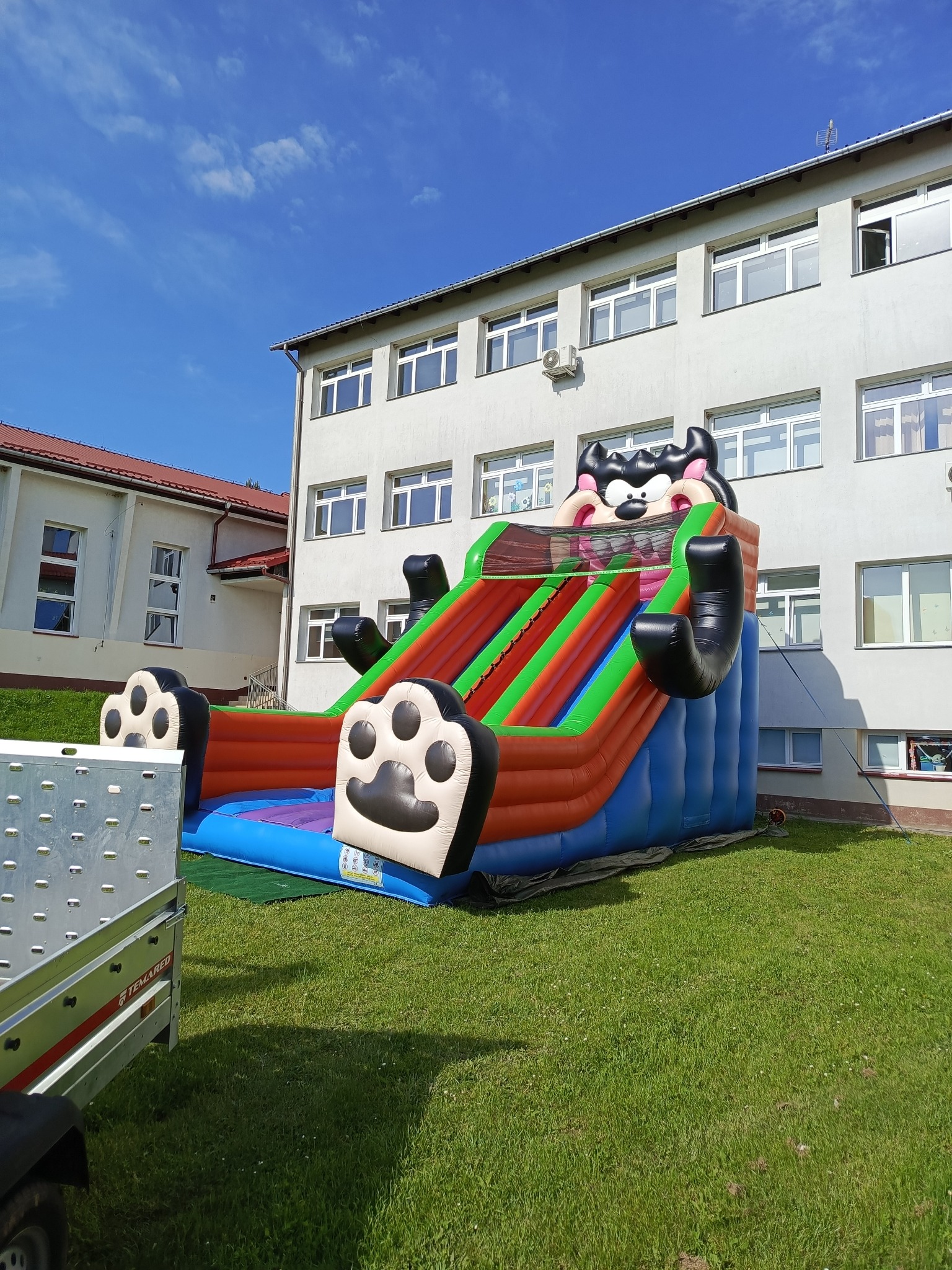 Kolorowa, dmuchana zjeżdżalnia z motywem kreskówkowym na trawie przed budynkiem. Widoczny fragment przyczepy transportowej.