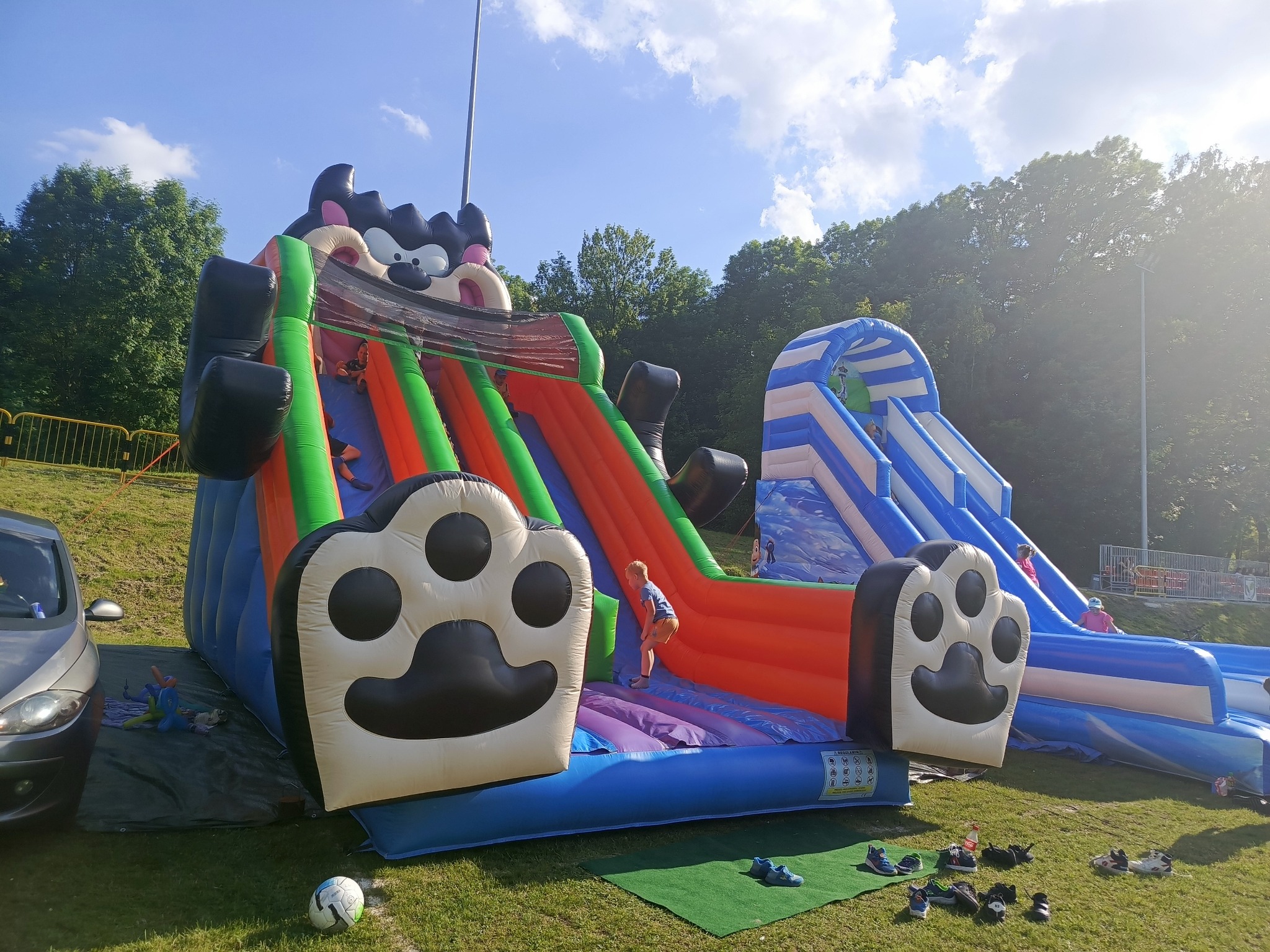 Kolorowy dmuchany plac zabaw z motywem zwierzęcym, dzieci w trakcie zabawy, buty pozostawione obok, w tle zieleń drzew i błękitne niebo. Słoneczny dzień.