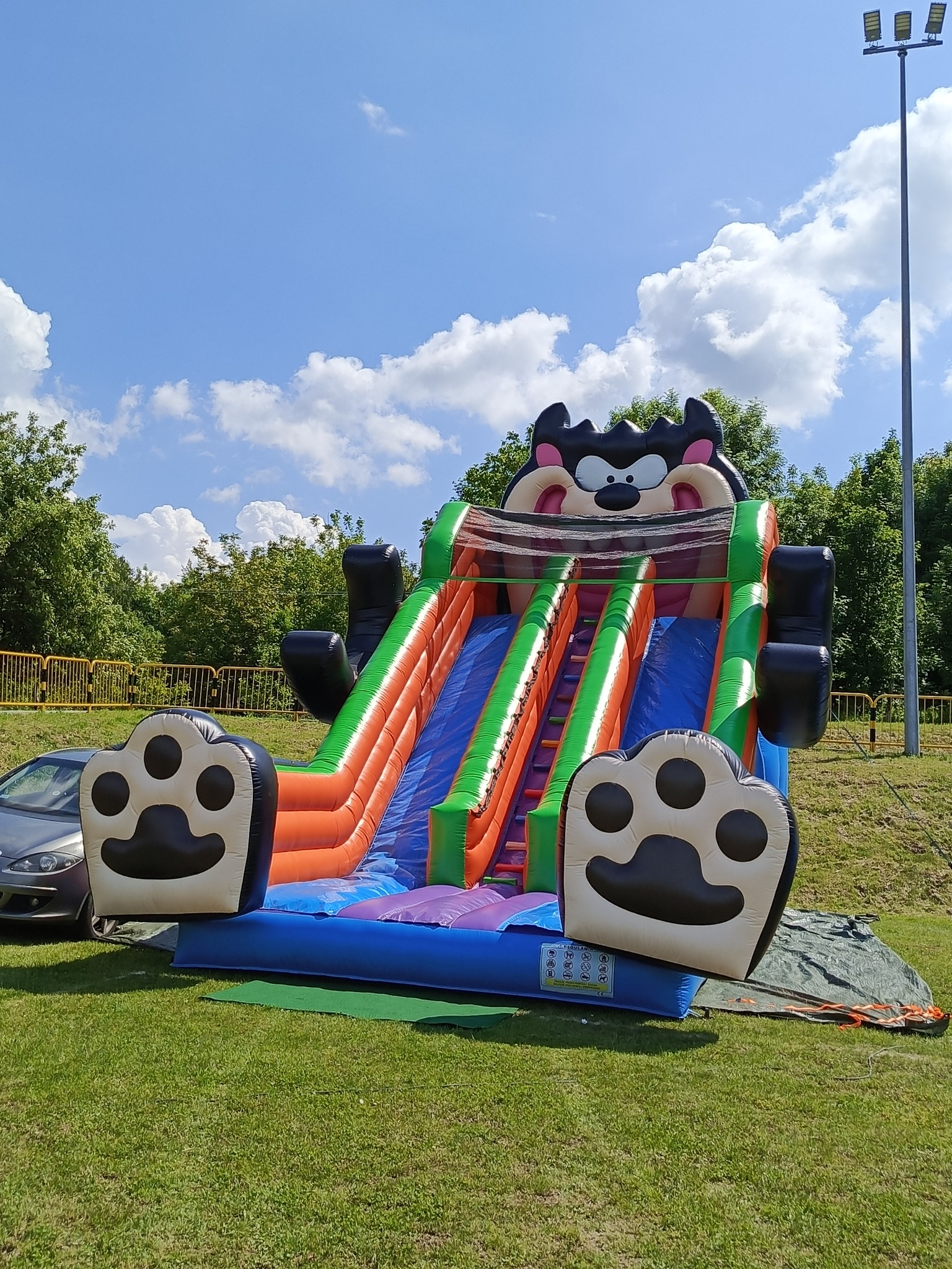 Kolorowa dmuchana zjeżdżalnia z motywem kreskówkowym na trawie, widok z dołu, błękitne niebo z chmurami w tle, częściowo widoczny samochód po lewej stronie.