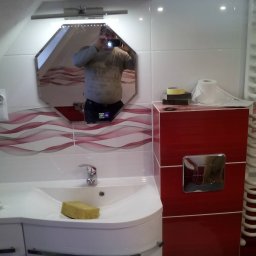 P.W. MIREX - Wykończona łazienka z umywalką, lustrem w kształcie ośmiokąta odbijającym fotografa, czerwonymi płytkami z falistym wzorem i grzejnikiem drabinkowym.