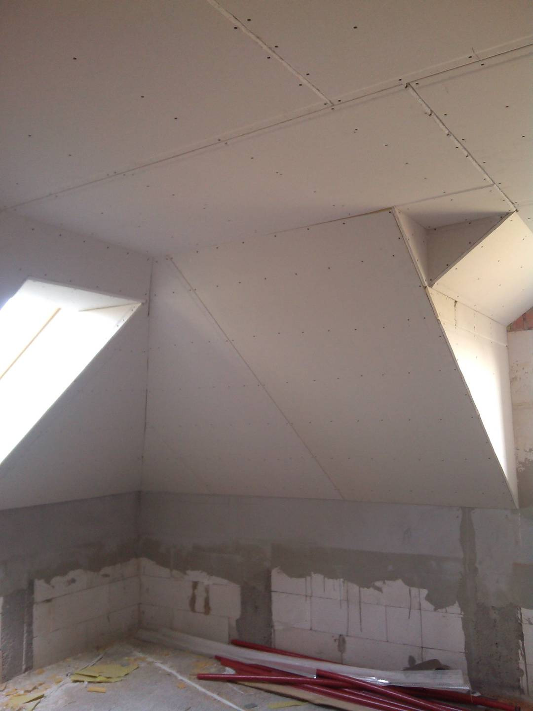 Poddasze w trakcie zabudowy płytami gipsowo-kartonowymi, widoczne okna dachowe i surowe ściany z betonu komórkowego, na podłodze rury PCV.