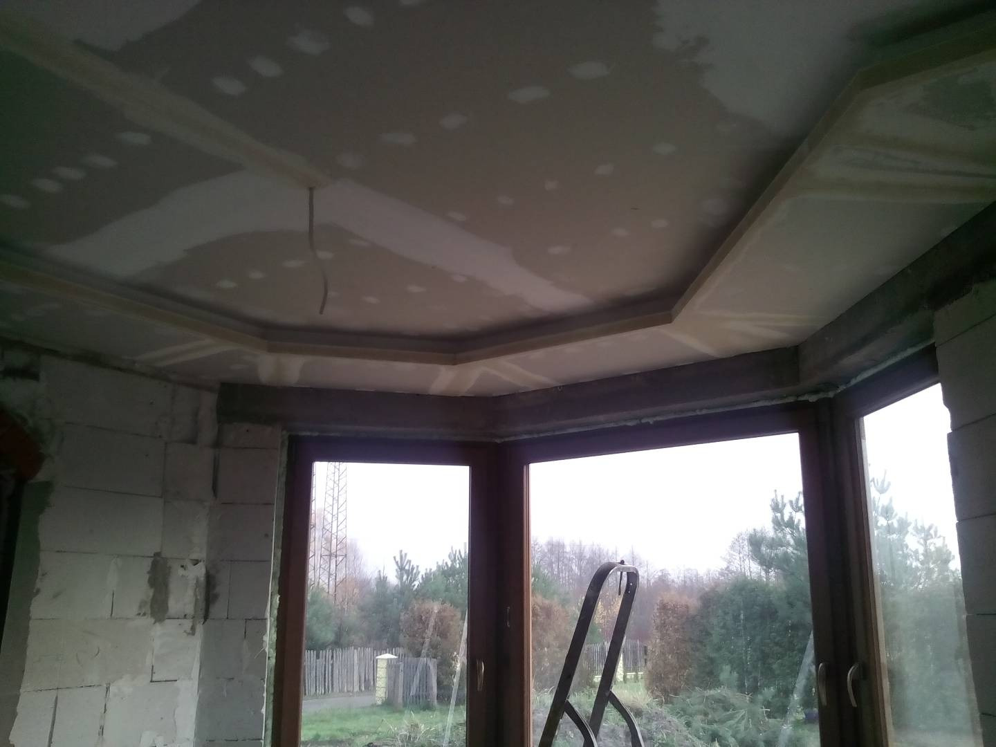 Montaż sufitu podwieszanego z płyt gipsowo-kartonowych z ozdobnym obramowaniem w narożniku pokoju, widok na okno z drabiną opartą o ramę.