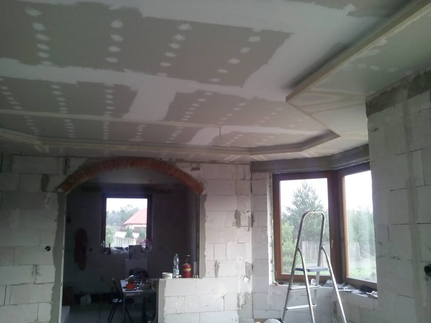 Wnętrze w trakcie prac wykończeniowych: sufit z płyt gipsowo-kartonowych ze szpachlowanymi łączeniami, murowany łuk z cegły nad oknem, okno z metalową drabiną opartą o parapet.