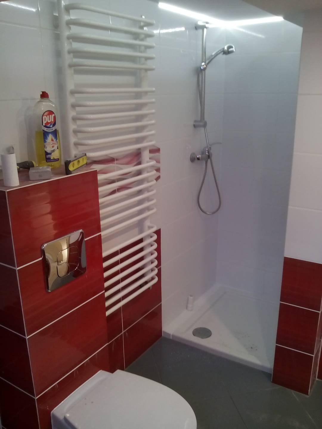 Wykończona łazienka z czerwono-białymi płytkami, białą kabiną prysznicową, grzejnikiem drabinkowym, i toaletą; na półce płyn do mycia naczyń Pur i poziomica.