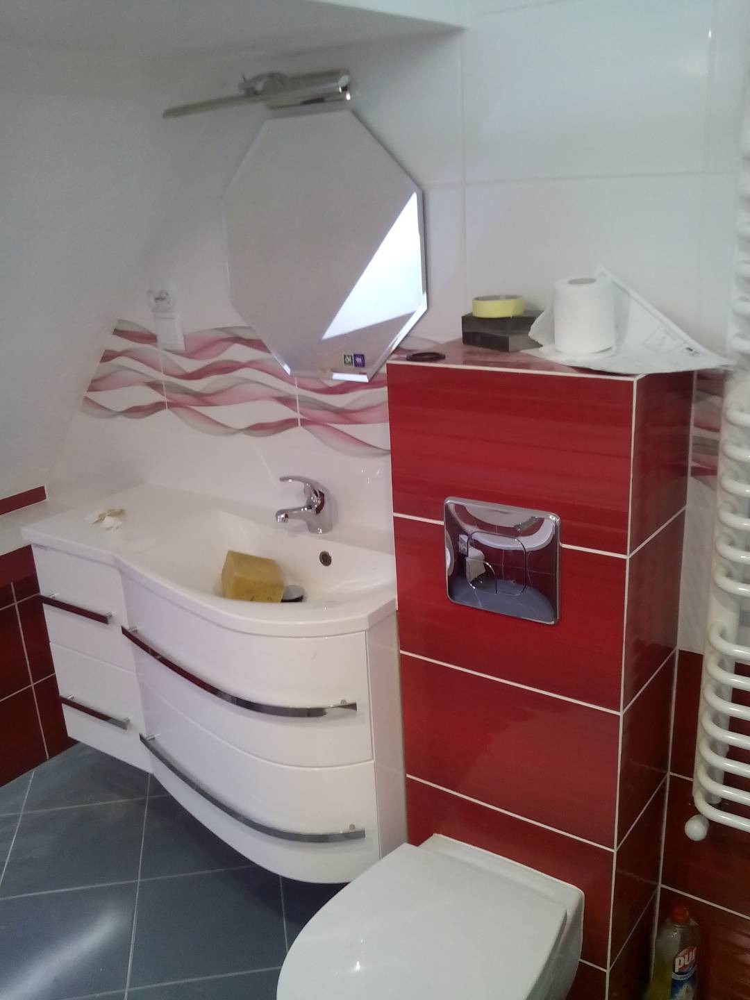 Ukończona łazienka z białą, zaokrągloną szafką pod umywalką, czerwonymi płytkami na ścianie, lustrem w kształcie ośmiokąta i toaletą. Na umywalce leży żółta gąbka.