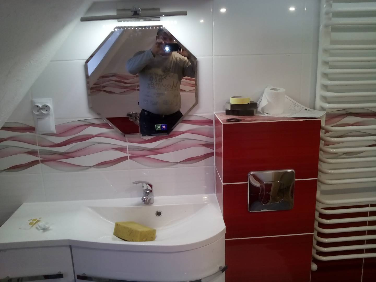 Wykończona łazienka z umywalką, lustrem w kształcie ośmiokąta odbijającym fotografa, czerwonymi płytkami z falistym wzorem i grzejnikiem drabinkowym.