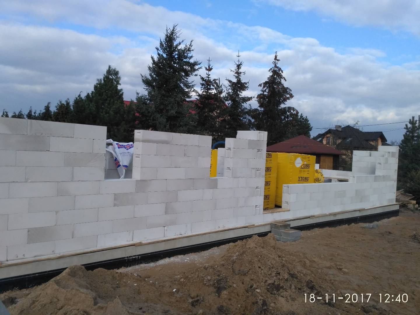 Wznoszenie ścian domu z białych bloczków Ytong, widoczne żółte opakowania bloczków, w tle drzewa i zabudowania.