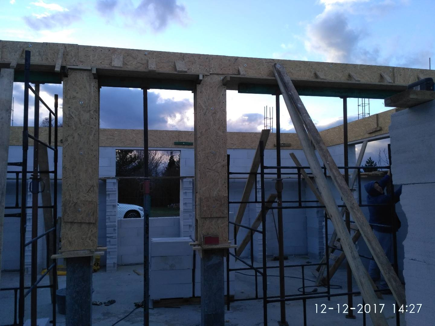 Budowa domu w trakcie: murowane ściany z betonu komórkowego, drewniane szalunki okienne, stalowe rusztowania, widoczny pracownik w kurtce.