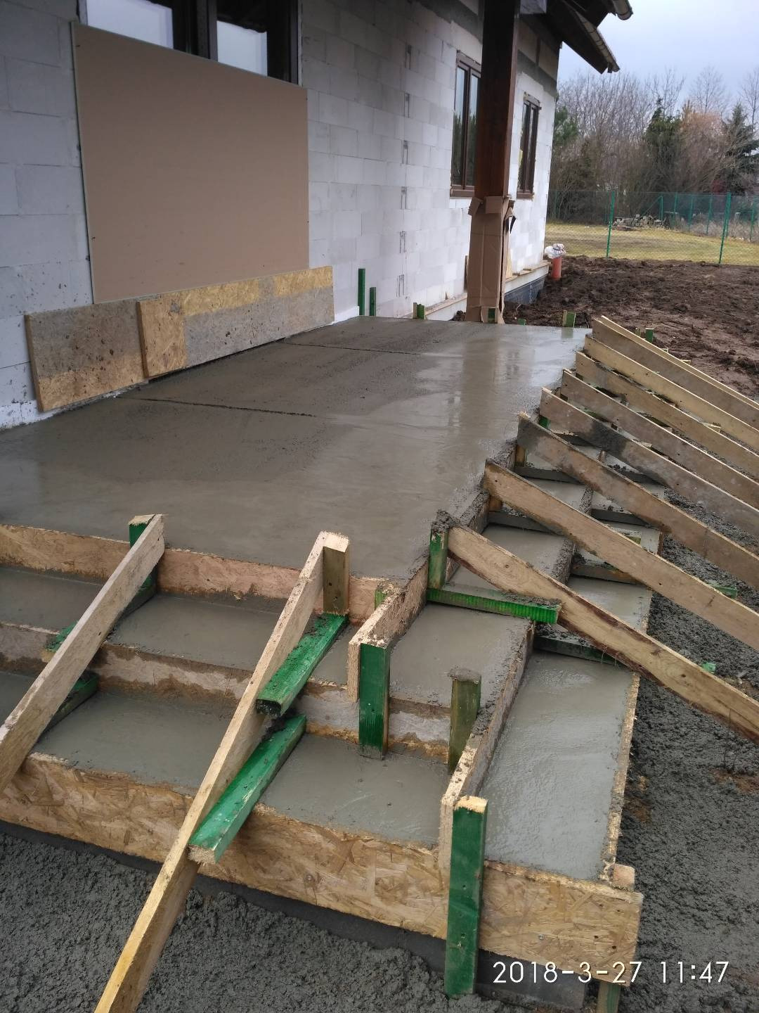 Wylewanie betonowej wylewki tarasu z drewnianym szalunkiem i zielonymi wspornikami, widoczny fragment ściany budynku z pustaków.