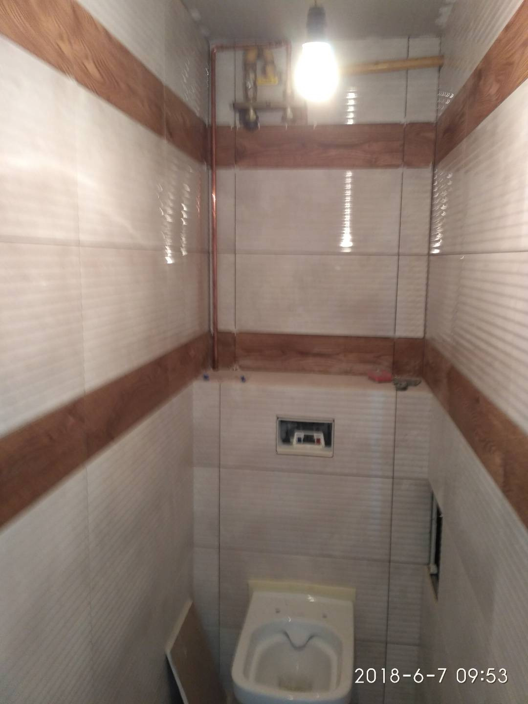 Wnętrze łazienki w trakcie wykańczania z białymi płytkami z drewnopodobnym pasem, widoczna toaleta i rury miedziane pod sufitem oraz otwór w ścianie.