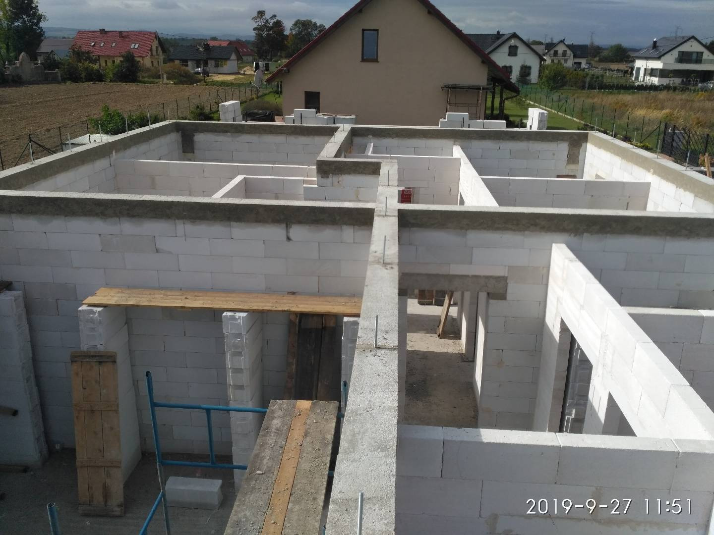 Wznoszenie ścian parteru domu z betonu komórkowego, widoczne szalunki nadproży okiennych i drzwiowych, w tle zabudowa jednorodzinna i pole uprawne.