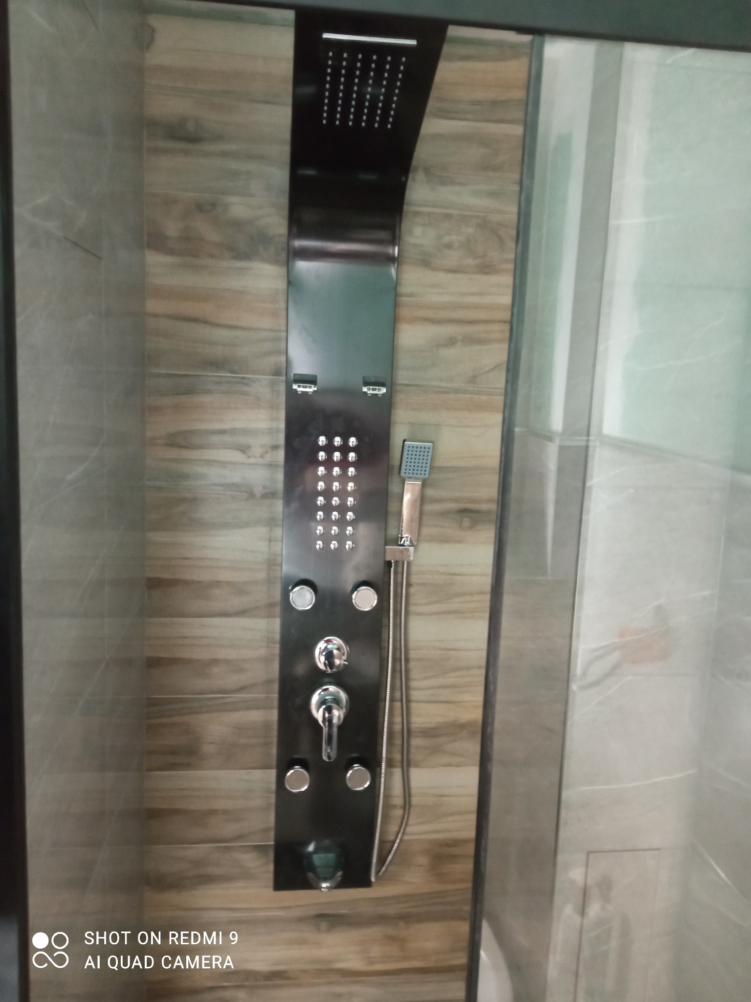 Czarny panel prysznicowy z deszczownicą i hydromasażem, zamontowany na ścianie wyłożonej płytkami imitującymi drewno w kabinie prysznicowej.