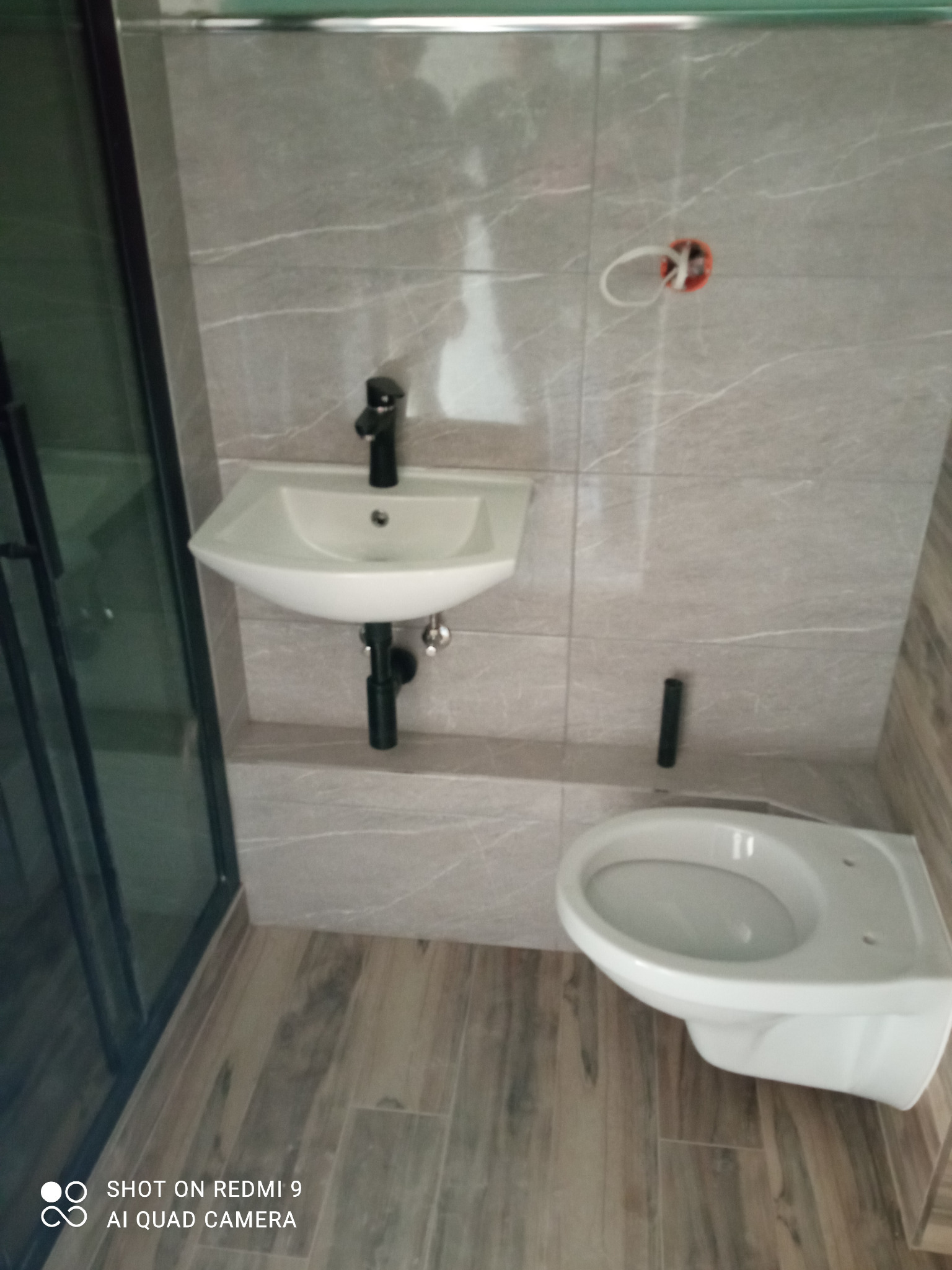 Nowoczesna łazienka z szarymi płytkami imitującymi marmur, podwieszana umywalka z czarną armaturą, wisząca toaleta i fragment kabiny prysznicowej z czarnymi profilami.