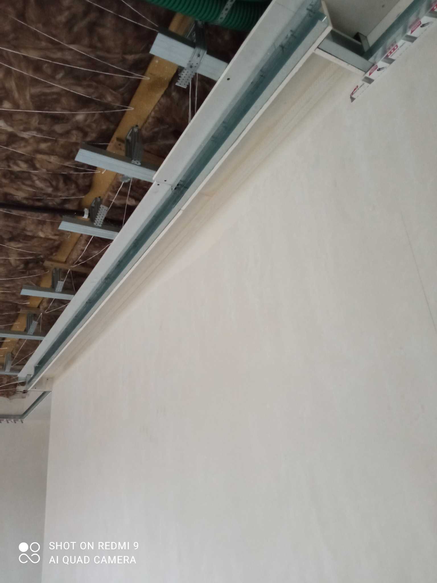 Sufit podwieszany w trakcie montażu, widoczne profile metalowe, izolacja z wełny mineralnej i fragment ściany pomalowanej na biało.