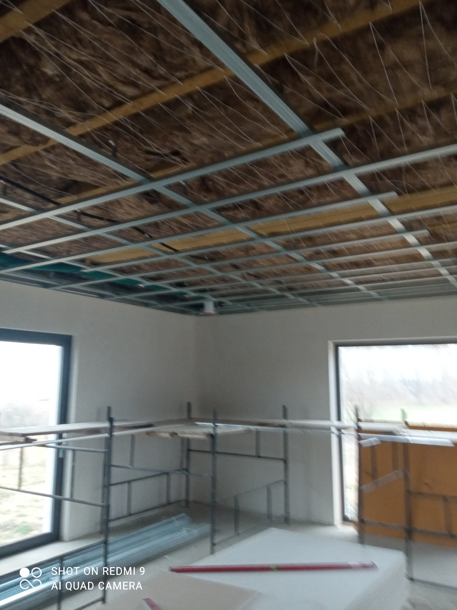 Sufit podwieszany w trakcie montażu: widoczna metalowa konstrukcja, izolacja termiczna i akustyczna, rusztowanie w pomieszczeniu z oknami.