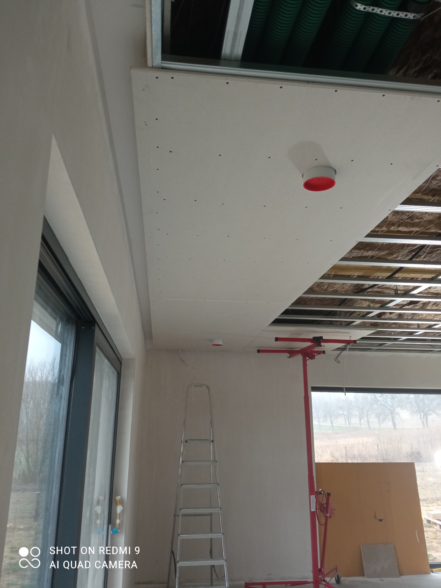 Montaż płyty gipsowo-kartonowej sufitu podwieszanego z widoczną konstrukcją metalową, ociepleniem i elementami instalacji, drabina aluminiowa i podnośnik do płyt.