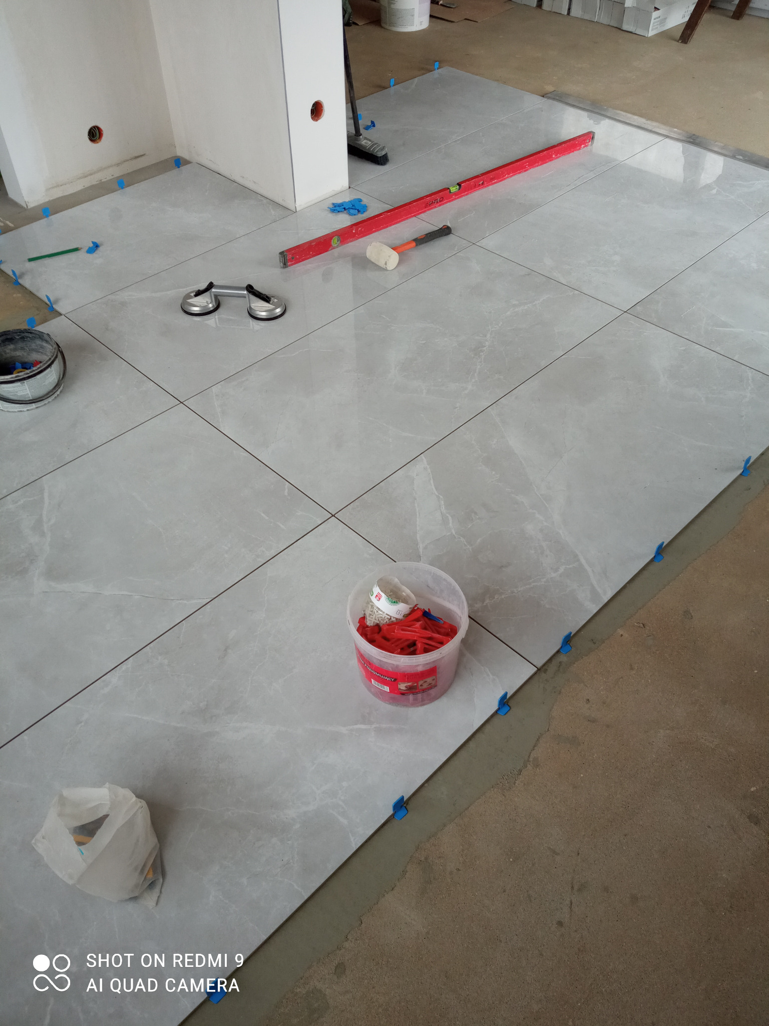 Układanie szarych płytek podłogowych z marmurkowym wzorem, widoczne narzędzia: poziomica, przyssawki do płytek, młotek gumowy, krzyżyki dystansowe w wiaderku i paczce.