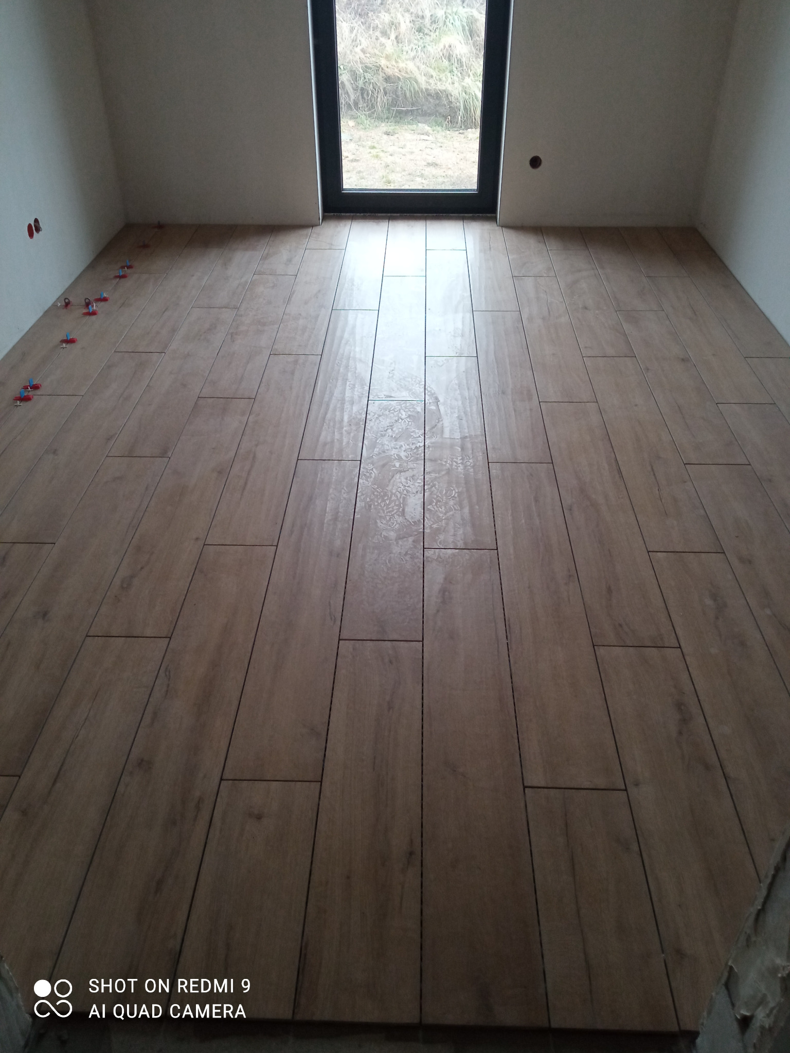 Świeżo ułożona podłoga z płytek imitujących drewno, z widocznymi klipsami poziomującymi wzdłuż ściany i oknem w tle.