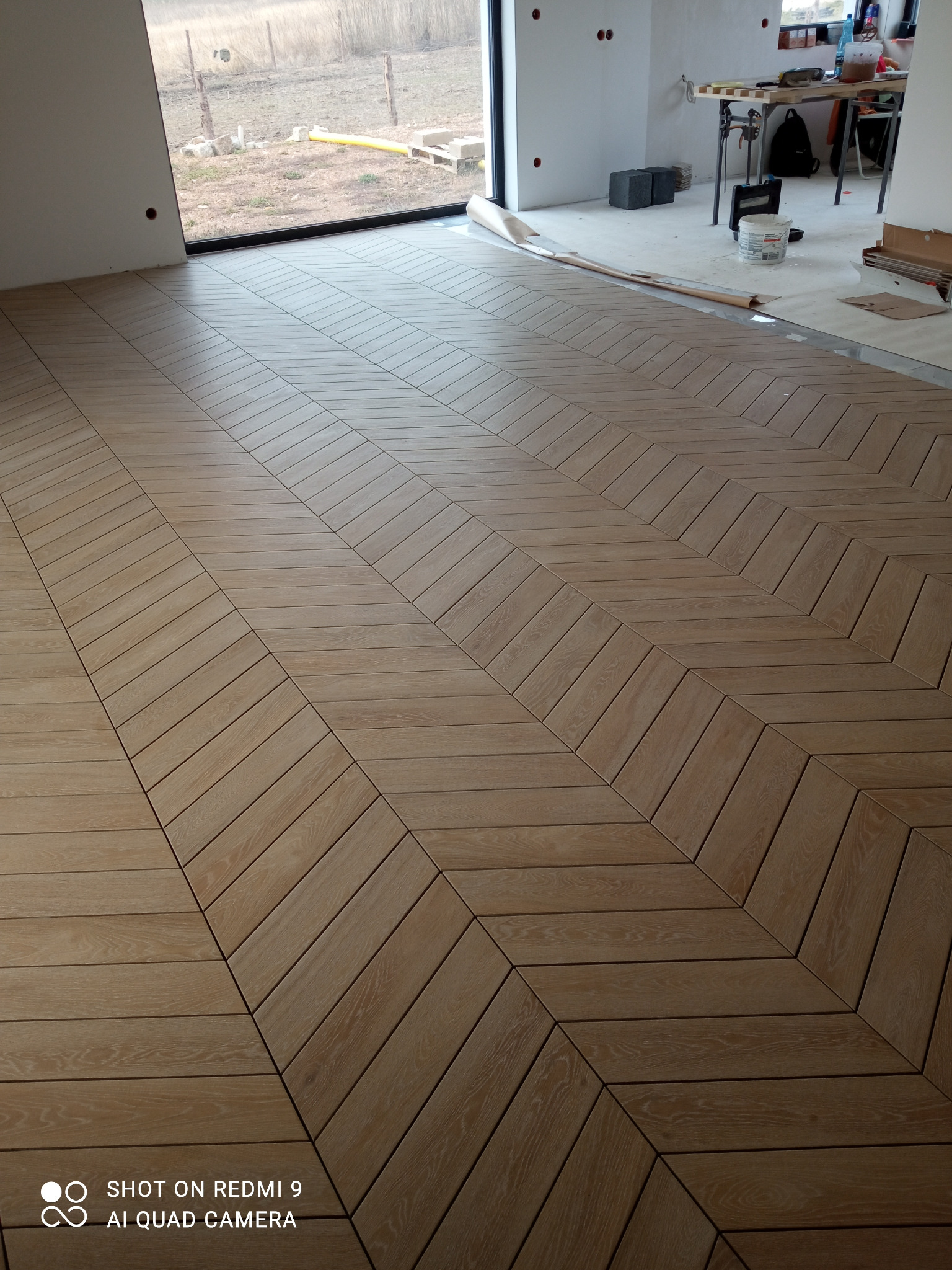 Ułożona podłoga z drewnopodobnych paneli w jodełkę, widoczne niedokończone prace wykończeniowe w tle, okno z widokiem na zewnątrz.