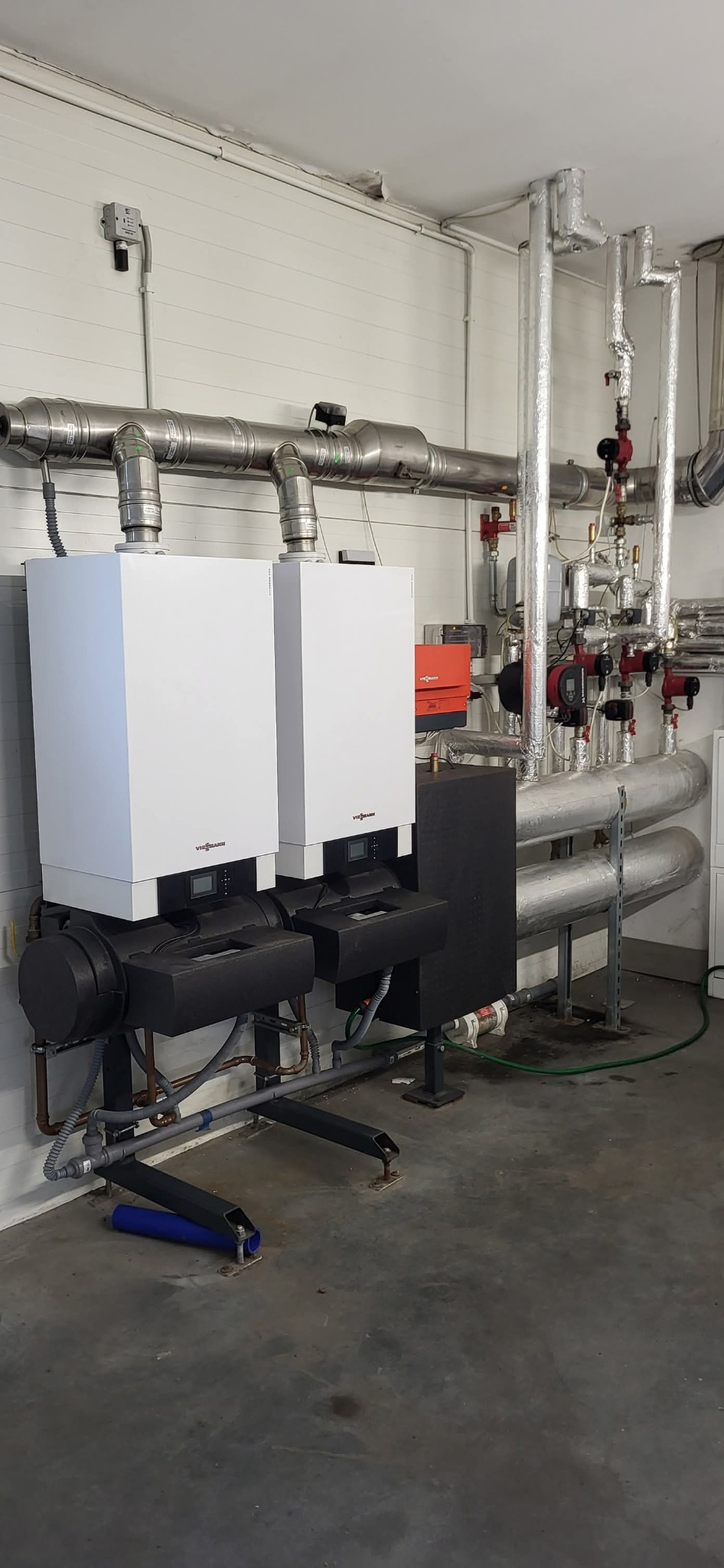 Dwa białe piece grzewcze Viessmann z czarnymi obudowami, połączone rurami, na tle białej ściany z instalacjami. Widoczne elementy instalacji grzewczej.
