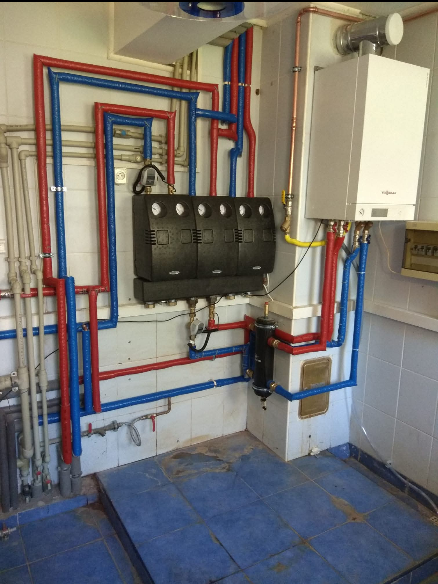 Złożona instalacja grzewcza z rurami w izolacji czerwonej i niebieskiej, kotłem Viessmann i czarnym rozdzielaczem, zamontowana w pomieszczeniu z niebieską podłogą.