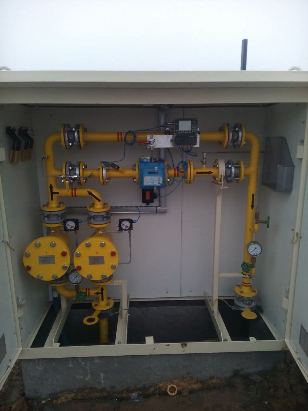 Żółta instalacja gazowa w szafce, z manometrami i zaworami. Widoczne rury, przyłącza i elementy sterujące. Całość w jasnej, metalowej obudowie.