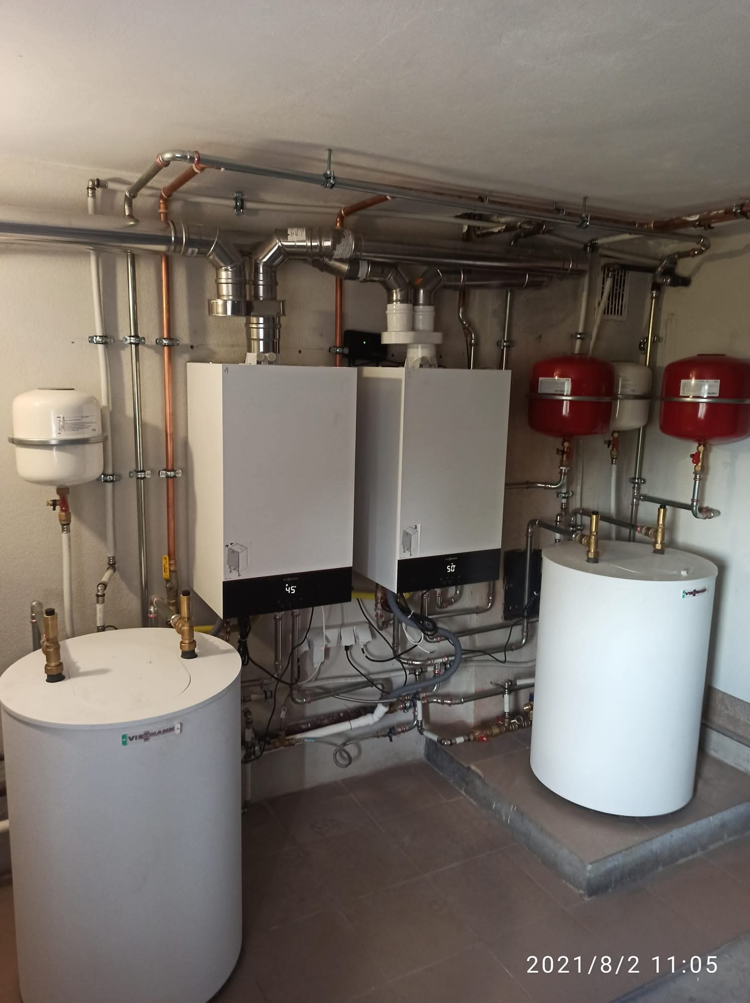 Dwa białe piece gazowe Viessmann z podłączonymi zbiornikami i instalacją hydrauliczną w pomieszczeniu kotłowni. Widoczne rury miedziane i stalowe.