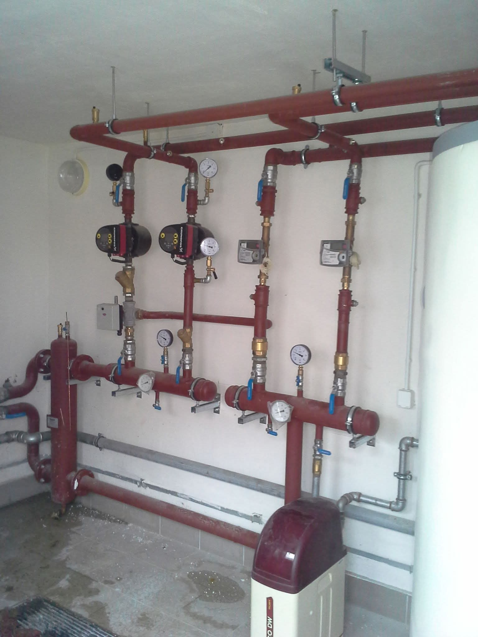 Złożona instalacja grzewcza z czerwonymi rurami, manometrami i pompami obiegowymi, zamontowana na białej ścianie w kotłowni. Widoczne elementy mocujące i zawory.