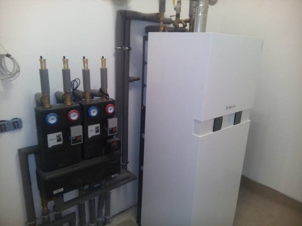 Nowoczesny piec grzewczy z osprzętem, widoczne manometry i rury. Biała obudowa pieca kontrastuje z ciemnymi elementami instalacji. Ujęcie z bliska, w pomieszczeniu.