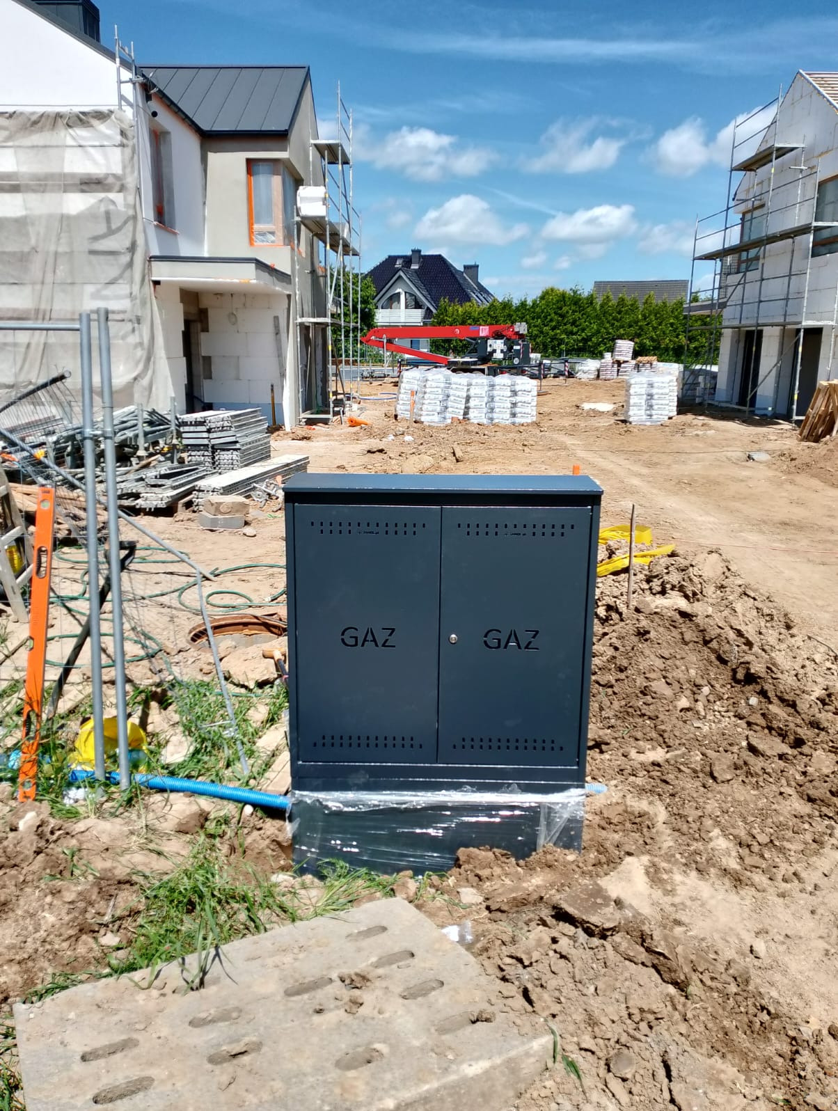 Szara szafka gazowa z napisem 'GAZ' na placu budowy, w tle domy w trakcie budowy i dźwig. Szafka zabezpieczona folią. Widoczne elementy instalacji.