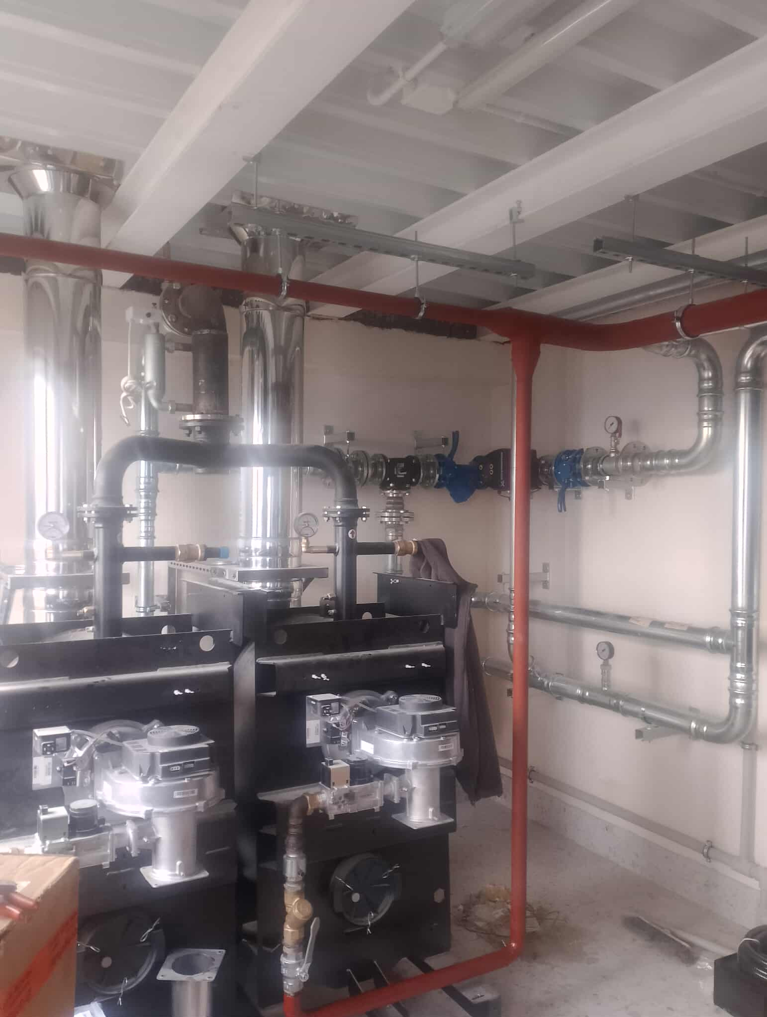 Złożona instalacja grzewcza z dwoma piecami, rurami w kolorze srebrnym i czerwonym, oraz elementami armatury. Widoczne manometry i zawory.