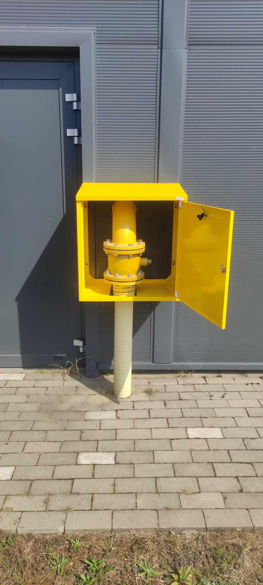 Żółta szafka gazowa z otwartymi drzwiczkami, zamontowana na szarej ścianie budynku. Widoczna instalacja gazowa wewnątrz szafki i chodnik z kostki brukowej.