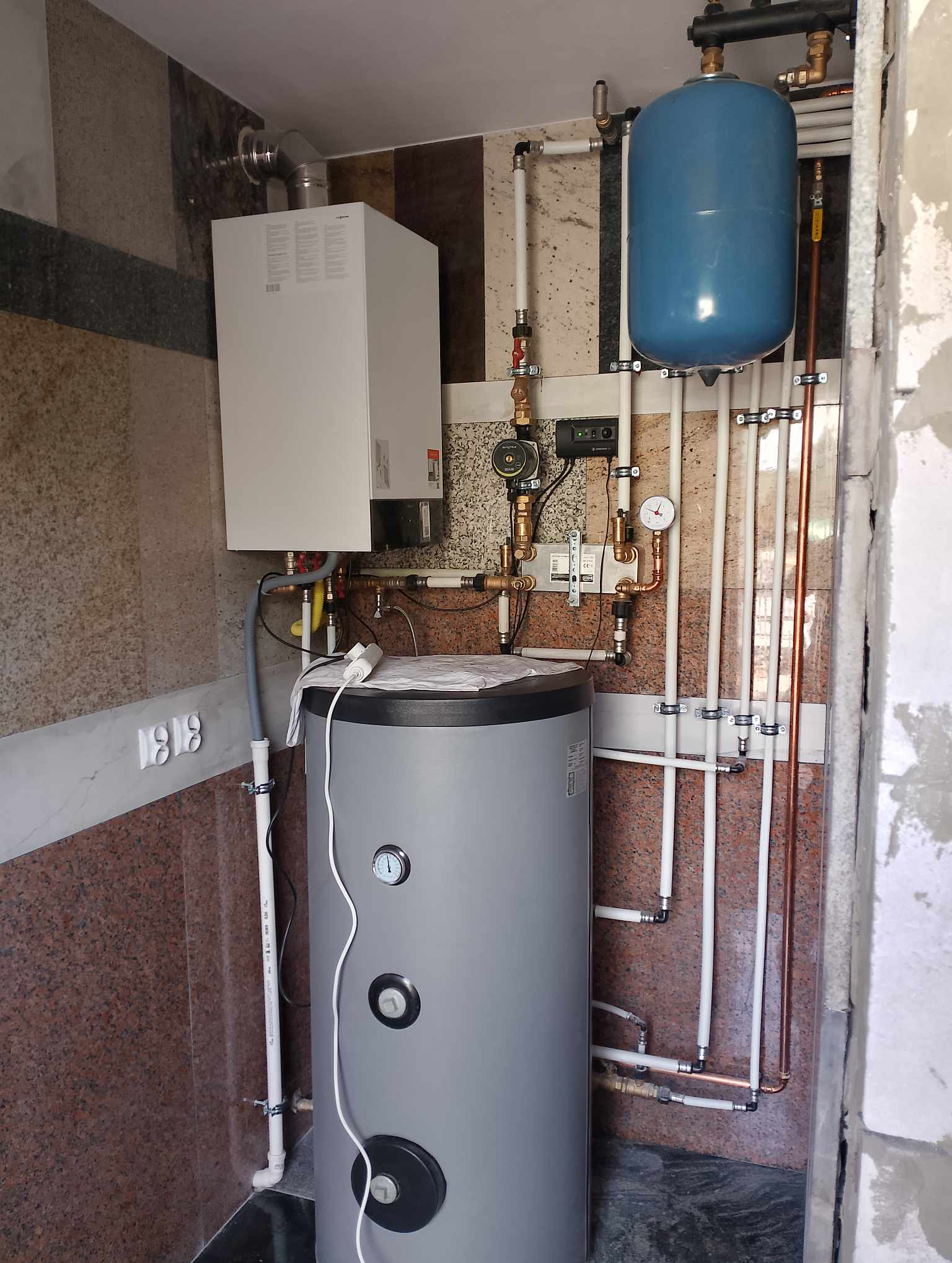 Kompaktowa kotłownia z piecem gazowym, zasobnikiem CWU i instalacją. Widoczne rury miedziane i plastikowe, manometry i armatura. Estetyczne wykończenie w płytkach.