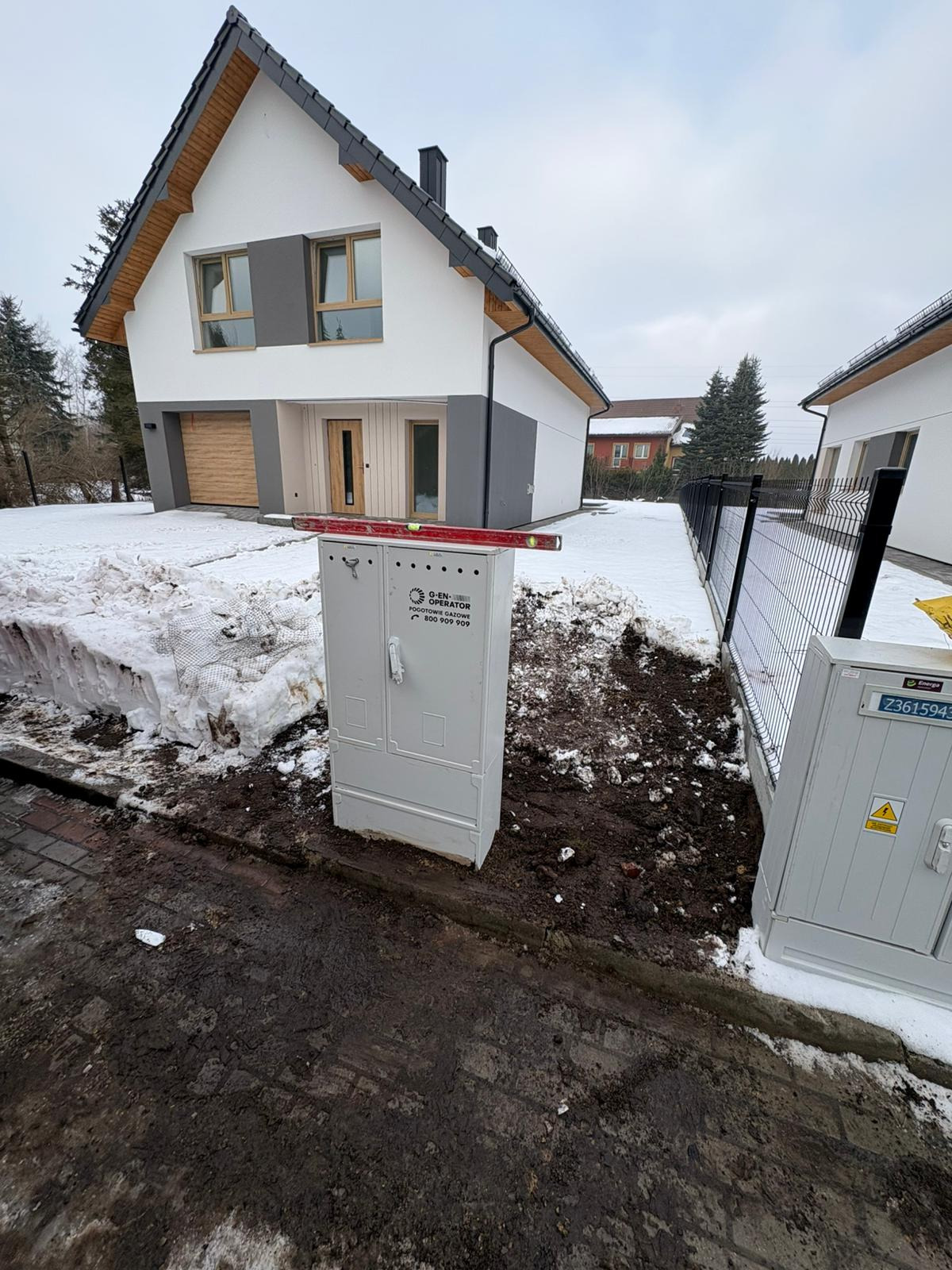 Szara skrzynka gazowa z poziomicą na górze, na tle nowoczesnego domu z drewnianymi akcentami i resztkami śniegu. Widoczne prace ziemne.