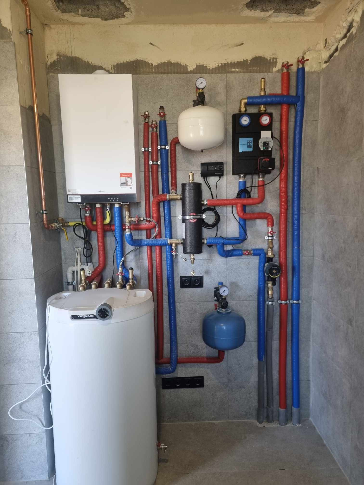 Złożona instalacja grzewcza Viessmann z kotłem, zasobnikiem i osprzętem, rury w izolacji czerwonej i niebieskiej, na tle szarej ściany z płytek. Widok z przodu.