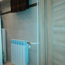 ProDom - Biały grzejnik panelowy z podłączeniem rur w łazience z beżowymi płytkami i brązowymi ręcznikami. Widoczne zawory i elementy instalacji.