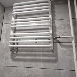 ProDom - Biały grzejnik drabinkowy zamontowany na szarej, wielkoformatowej płytce ściennej, z widocznym zaworem termostatycznym i rurami instalacji grzewczej.