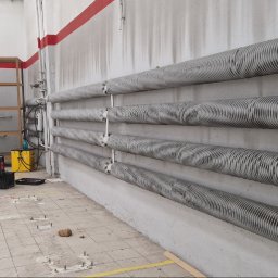 ProDom - Pięć poziomych, ożebrowanych rur instalacji centralnego ogrzewania na betonowej ścianie z czerwonym pasem. W tle regały i wózek transportowy w pomieszczeniu technicznym.