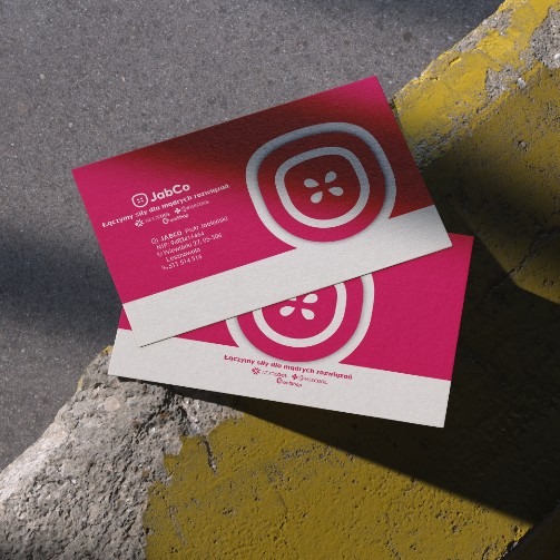 Dwie różowe wizytówki firmy JabCo z białym logo stylizowanym na guzik, leżące na szarym betonie z żółtymi pasami. Kontrastowe kolory i ostre cienie.