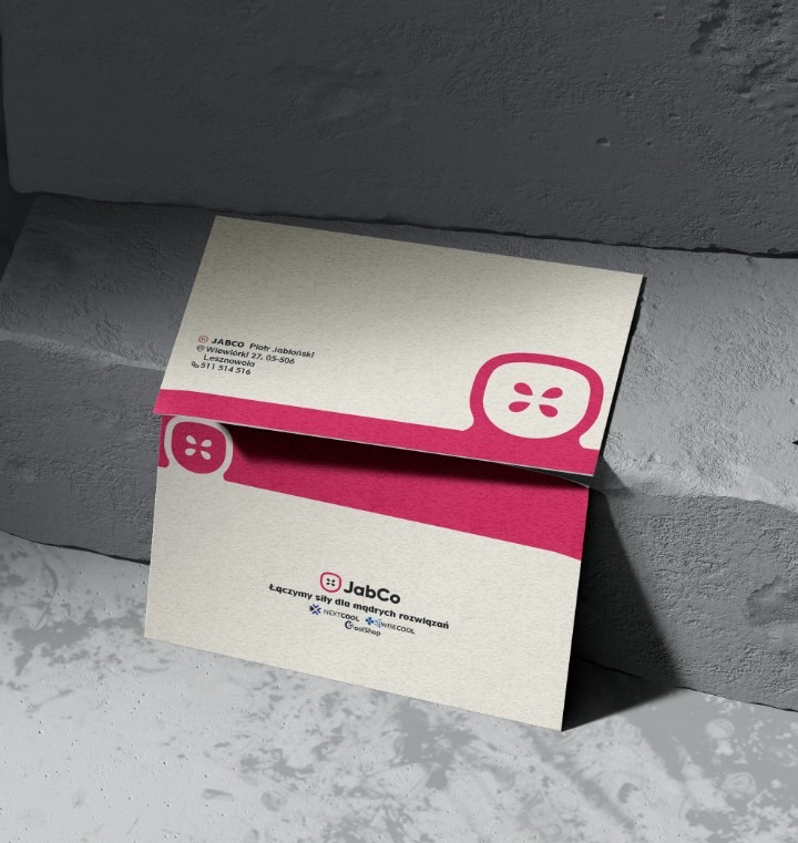 Elegancka wizytówka firmy JabCo w odcieniach bieli i fuksji, z nowoczesnym logo i hasłem na teksturowanym papierze, oparta o szarą, betonową ścianę.