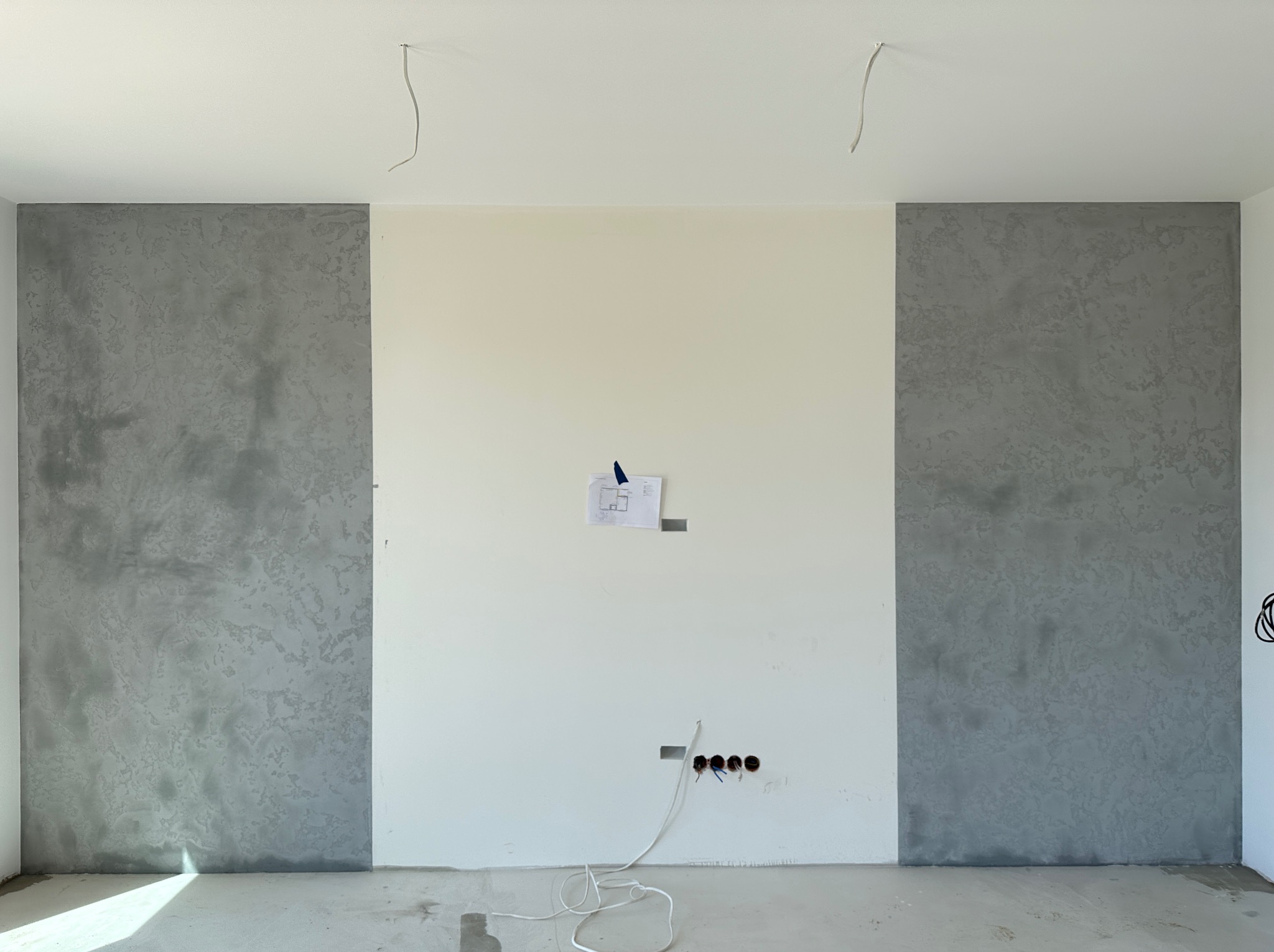 Ściana z betonem architektonicznym w Nowym Mieście Lubawskim. Szare panele po bokach, biała ściana pośrodku z planem i gniazdkami. Wystające kable.