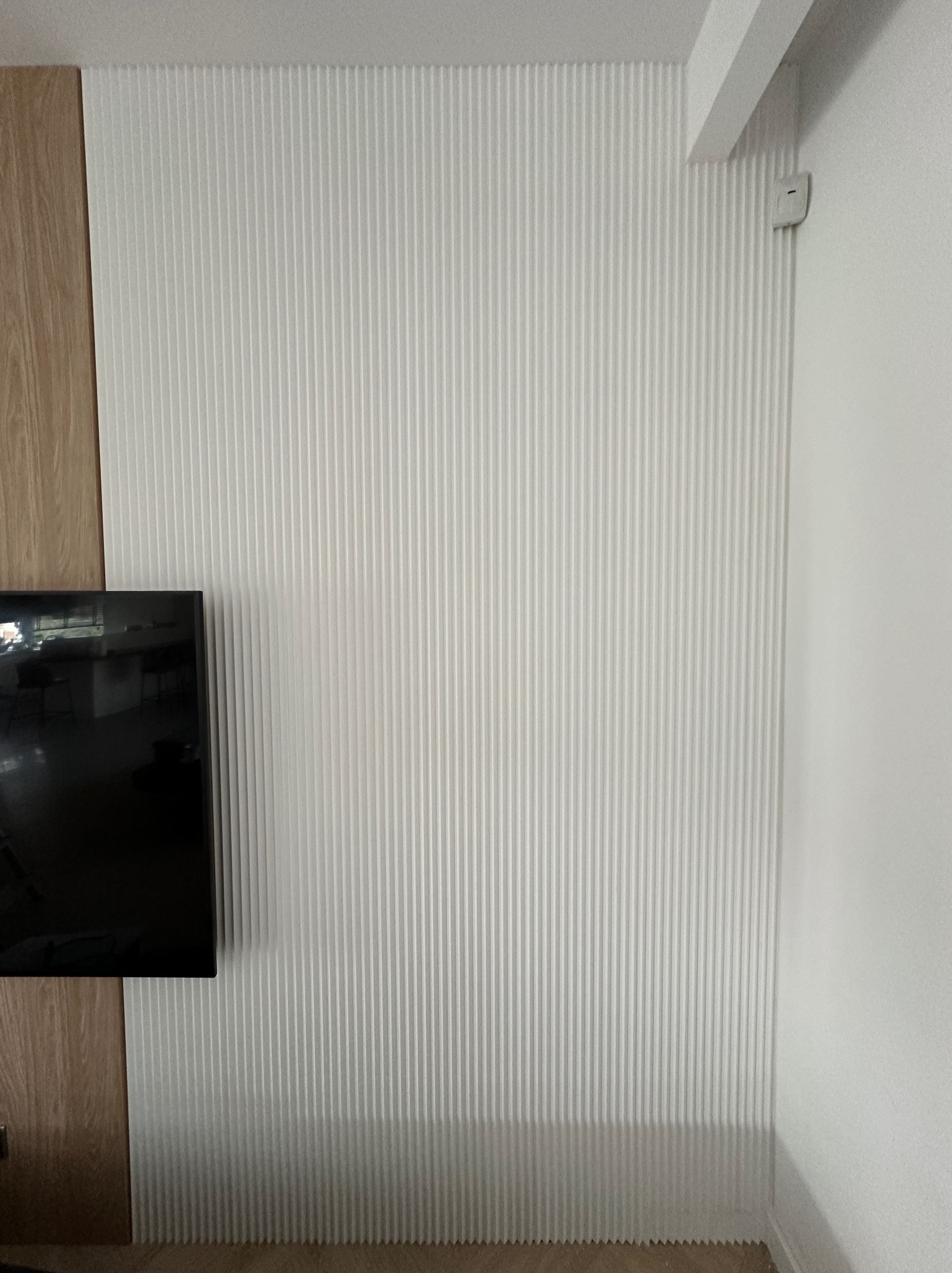 Biała, pionowo żłobkowana sztukateria ścienna, częściowo zakryta przez drewnianą okładzinę i telewizor. Widoczny fragment podłogi i narożnik ściany.