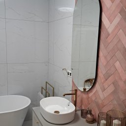 Park 82 - Nowoczesna łazienka z białą armaturą, różową ścianą w jodełkę i owalnym lustrem. Umywalka nablatowa, złota bateria i dekoracje dodają elegancji.