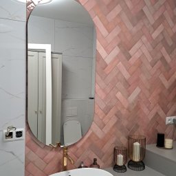 Park 82 - Nowoczesna łazienka z umywalką nablatową, złotą armaturą i różową ścianą z płytek ułożonych w jodełkę. W lustrze odbija się toaleta i szafka. Widoczne gniazdko elektryczne.