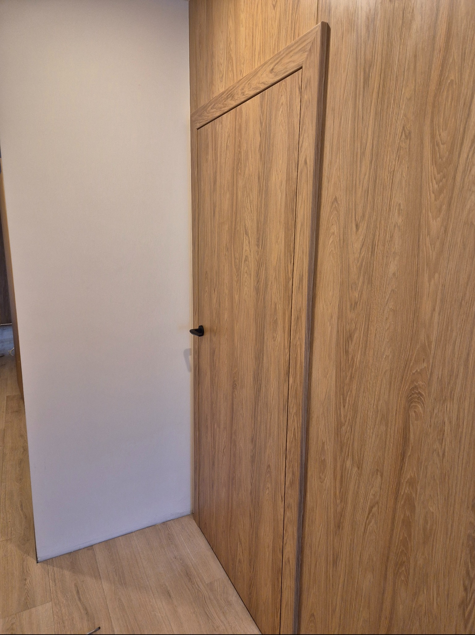 Zamknięte, drewniane drzwi z czarną klamką, wkomponowane w drewnianą ścianę, przylegające do białej ściany. Widoczny fragment podłogi z paneli.