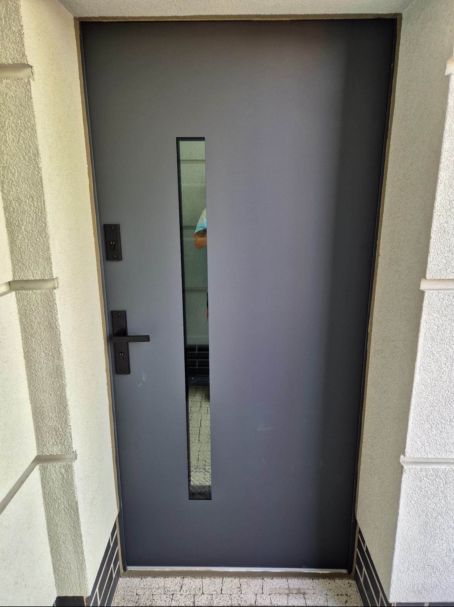 Nowoczesne, grafitowe drzwi wejściowe z wąskim przeszkleniem, zamontowane w jasnej elewacji z dekoracyjnym tynkiem i kamienną podmurówką. Minimalistyczny design.